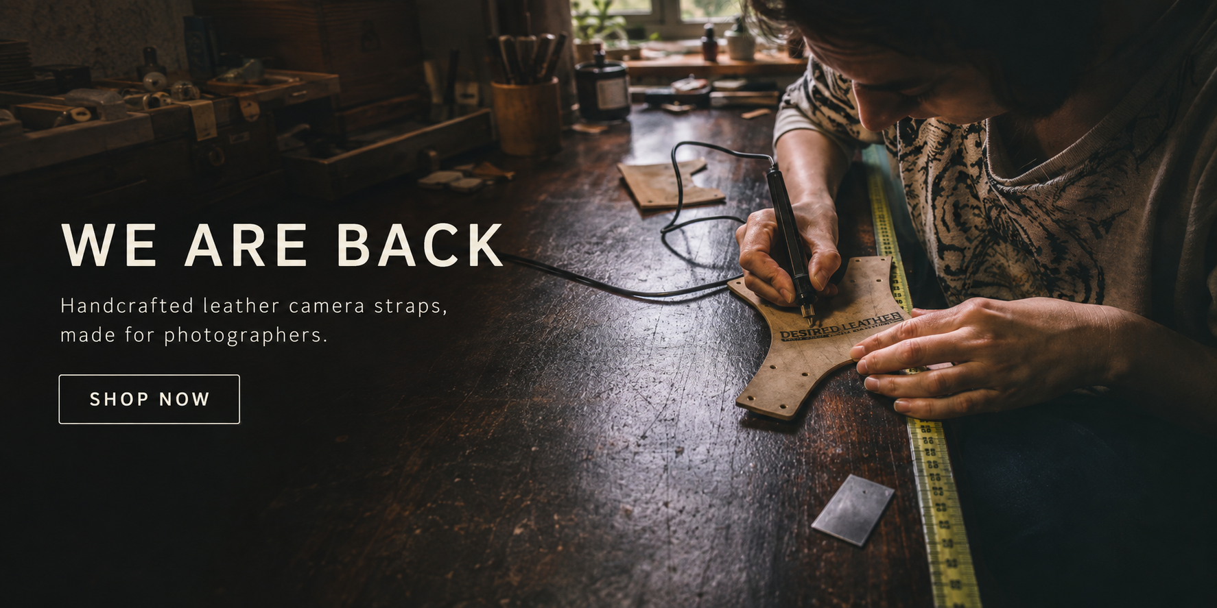 Artisan handcrafting a leather camera strap in the Desired Leather workshop beside a “We Are Back” announcement.